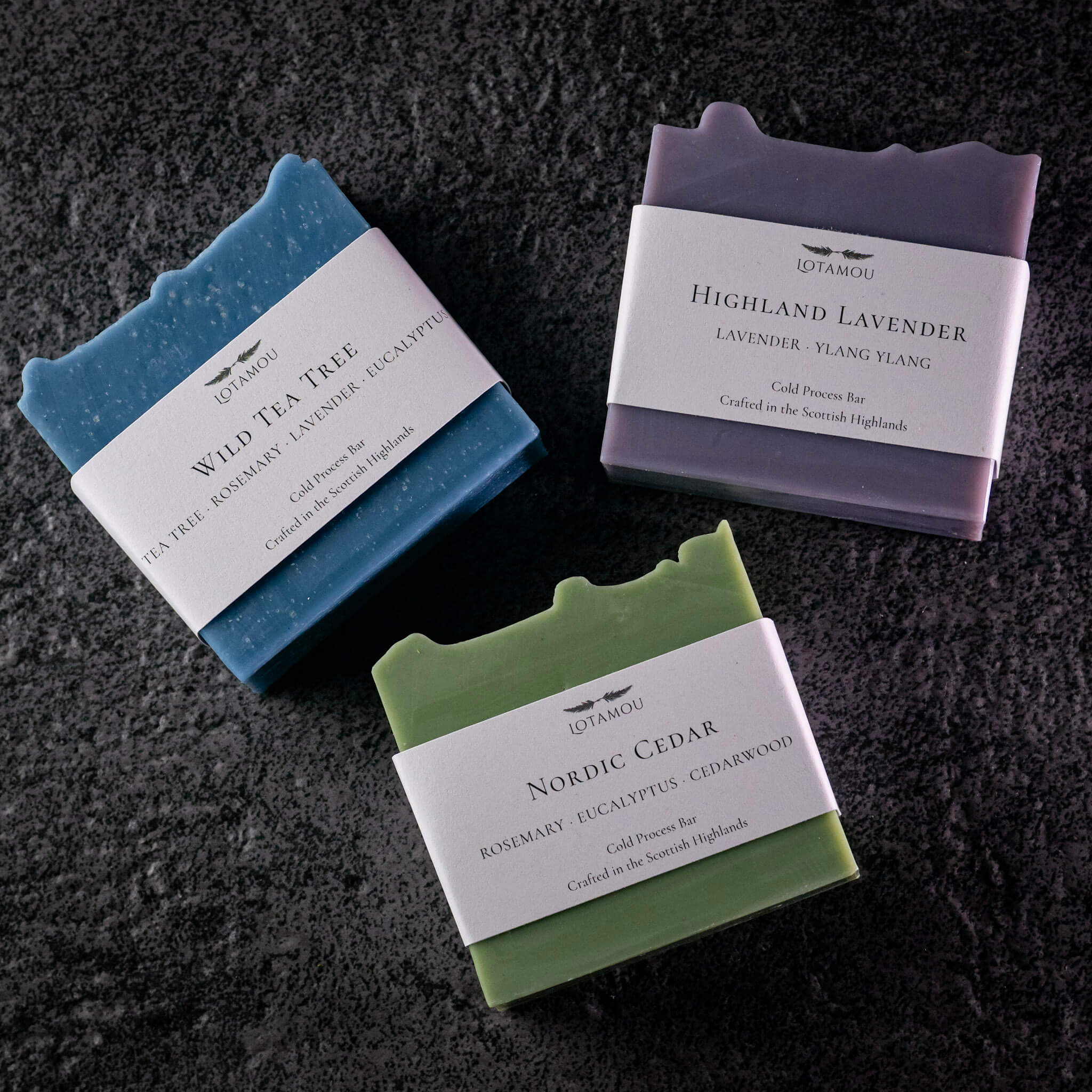 Three artisan cold-pressed soap bars from IDATABOU arranged on a dark textured background. The soaps are in blue, green, and purple colors, each with a white paper label showing the scent names: Wild Tea Tree, Nordic Cedar, and Highland Lavender.