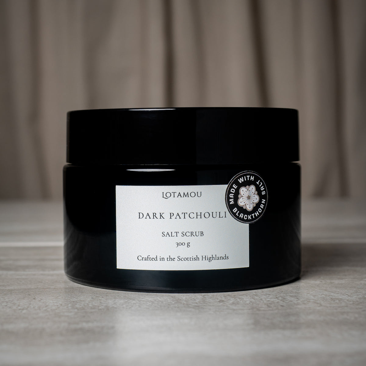 A black plastic jar of Lotamou Dark Patchouli Salt Scrub with a white label, set against a neutral gray textured surface and draped fabric background.