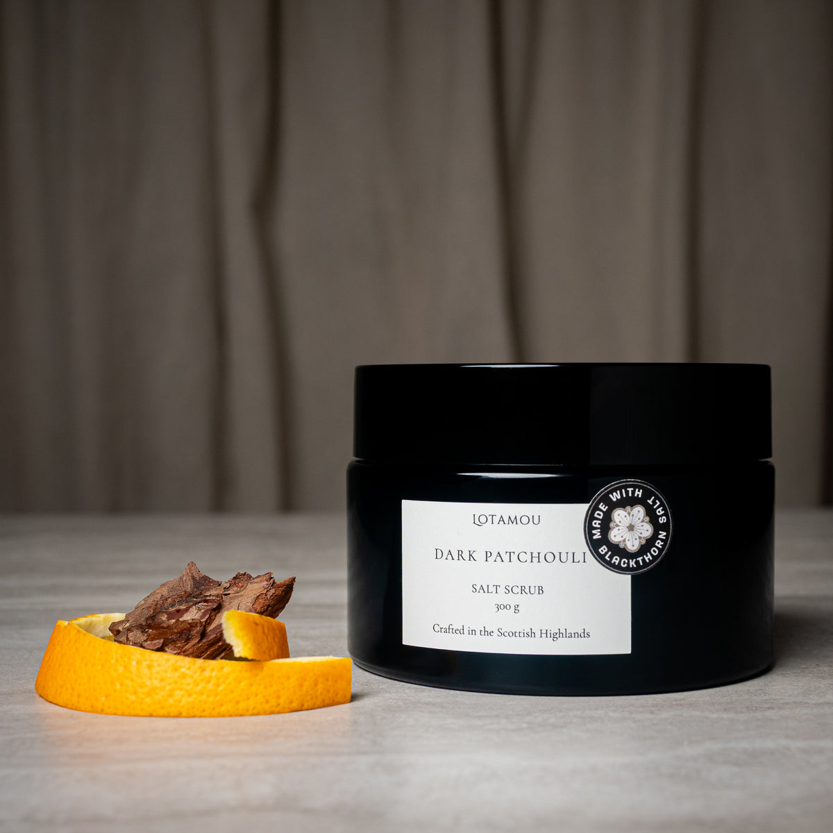 A black jar of LTAMOU Dark Patchouli Salt Scrub sits on a light gray surface with a dried patchouli herb piece and an orange slice beside it. The background is a soft-focus neutral brown fabric.