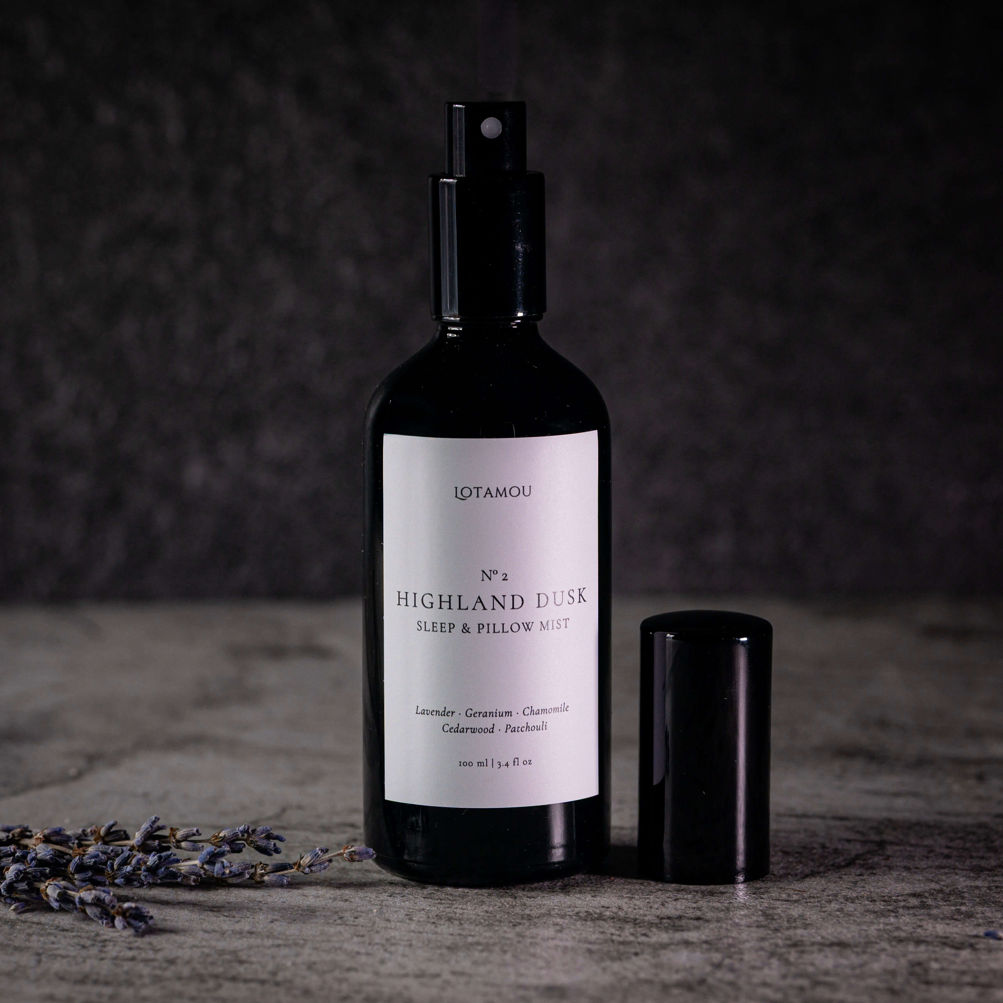 A dark glass spray bottle labeled 'IDTANOU No. 3 Highland Dusk Sleep & Pillow Mist' stands on a textured gray surface with its black cap beside it and dried lavender sprigs in the foreground against a dark gray background.