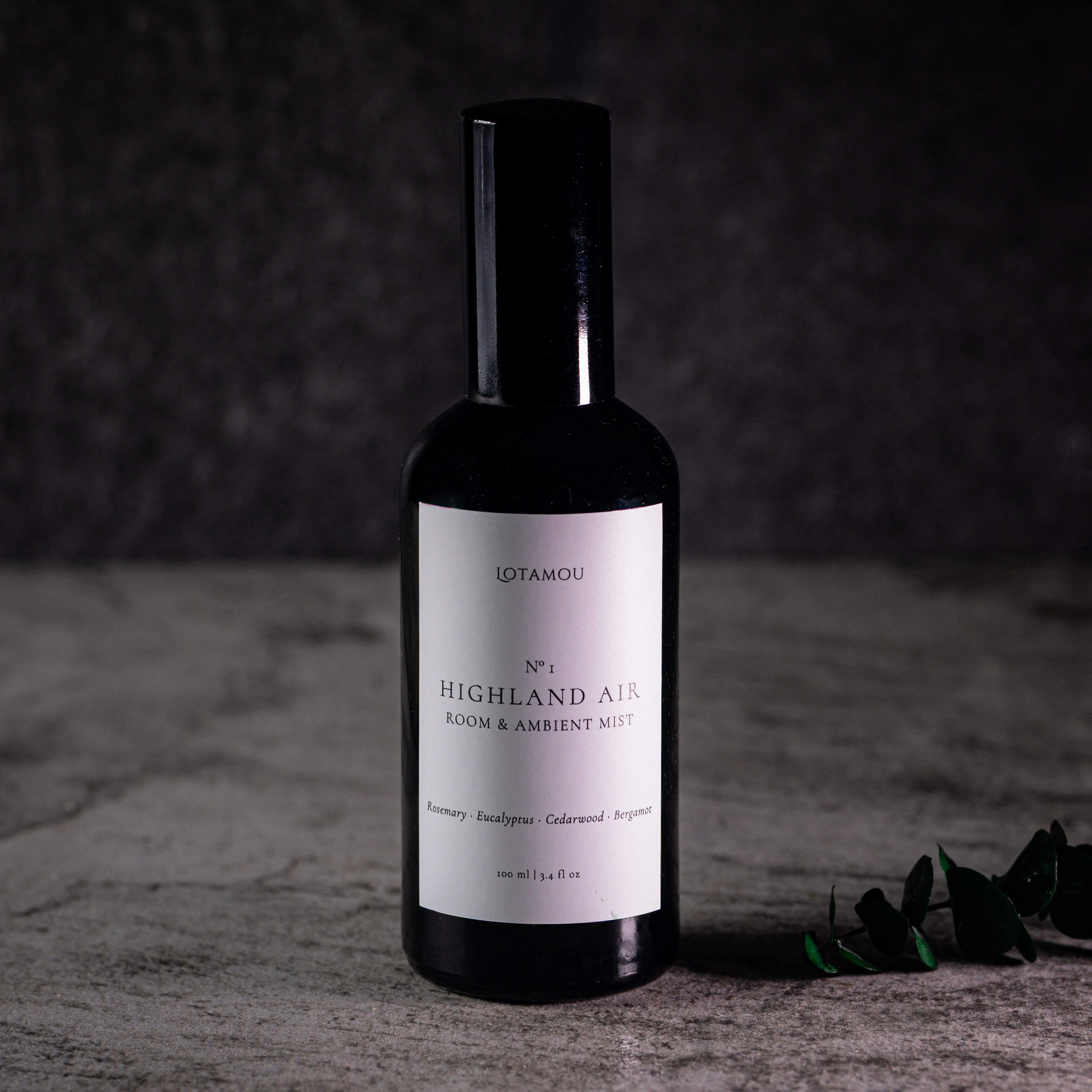 A dark glass bottle of Lotamou 'Highland Air' room and ambient mist spray, featuring a white label and a black spray cap, set against a dark textured background with a small green sprig on the side.