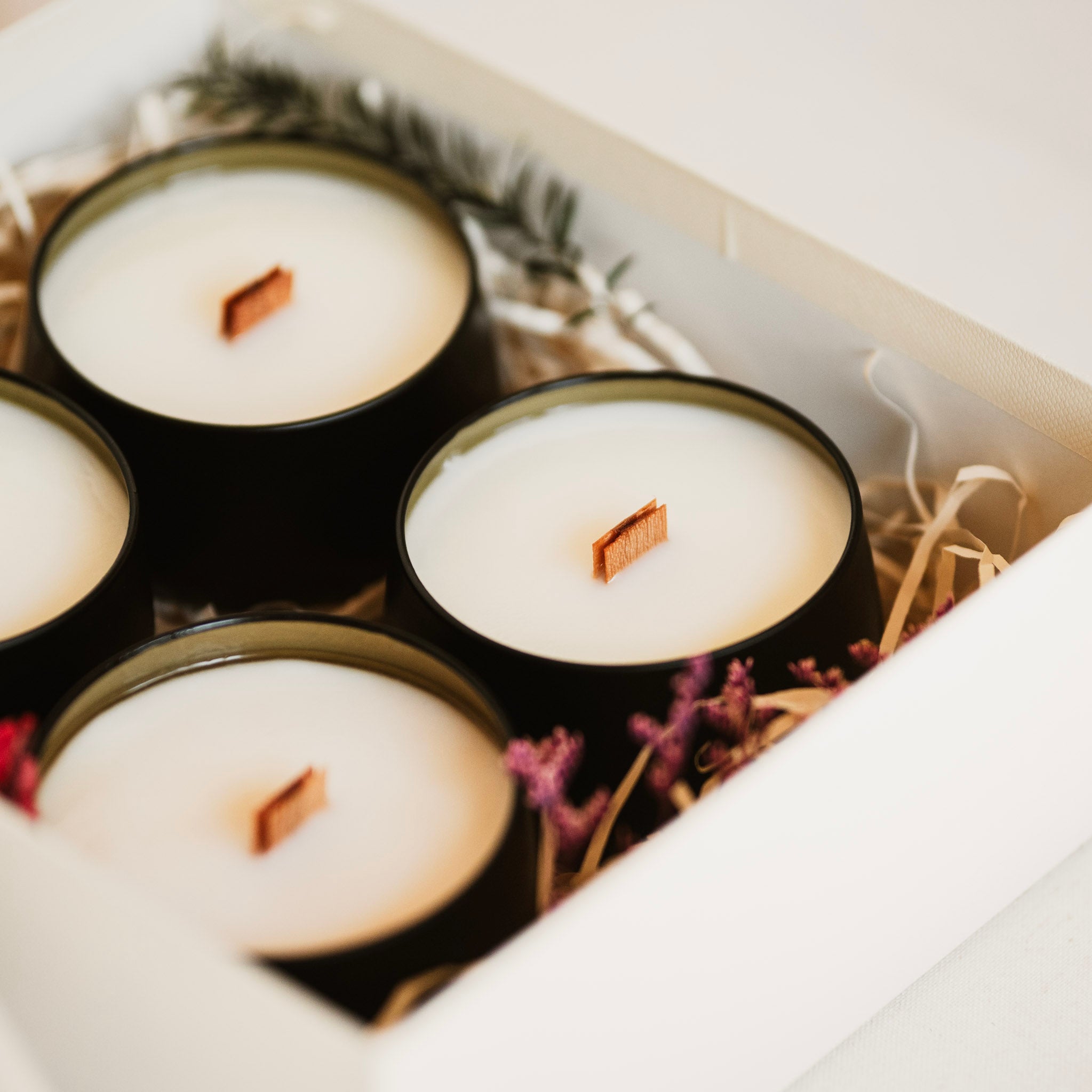 Four black candles with wooden wicks in a decorative box with straw and flowers.