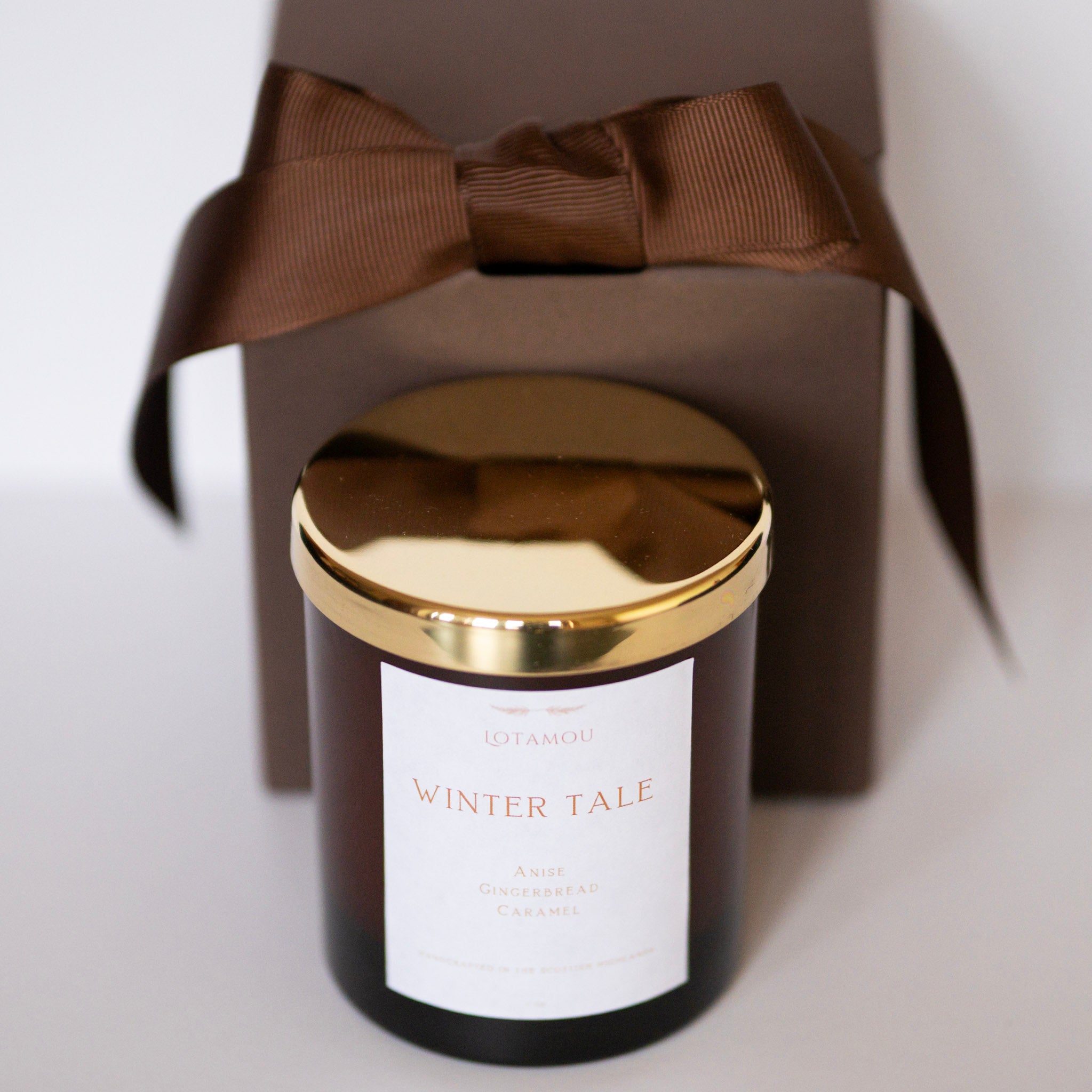 Candle with gold lid and 'Winter Tale' label in front of a brown gift box with a bow.