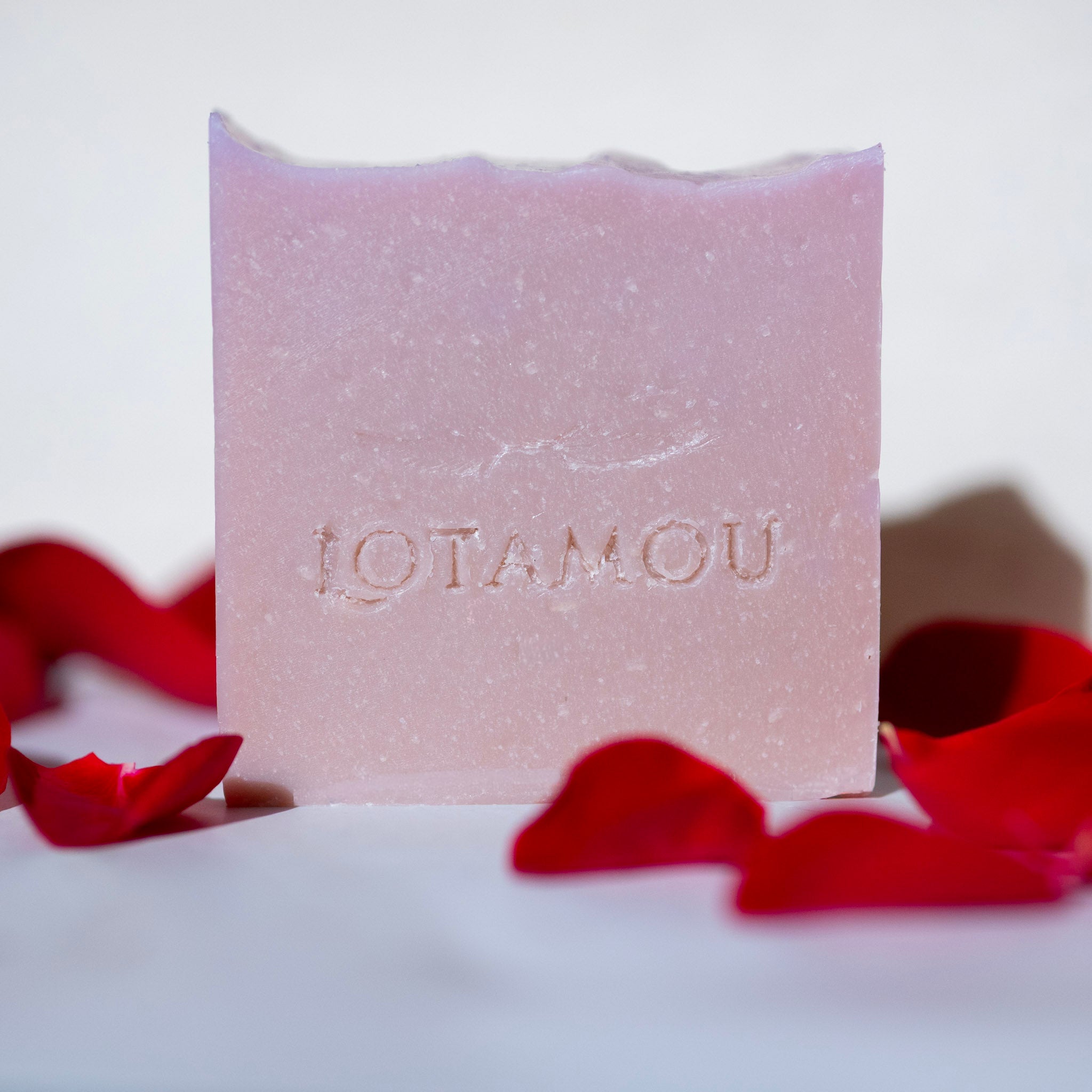 Bar of soap with 'LOTAMOU' branding surrounded by red rose petals on a white background
