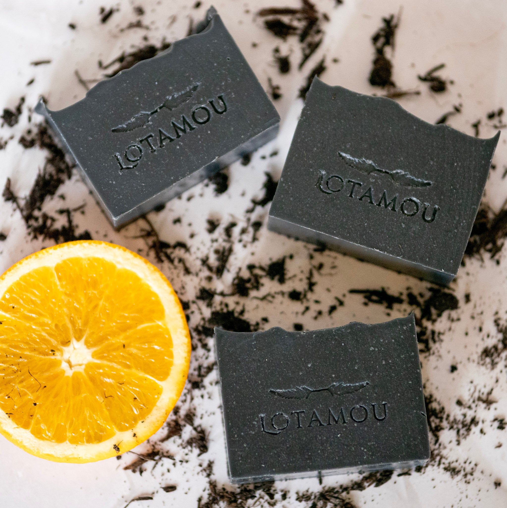 Three bars of Lotamou charcoal soap with a lemon on a textured surface.
