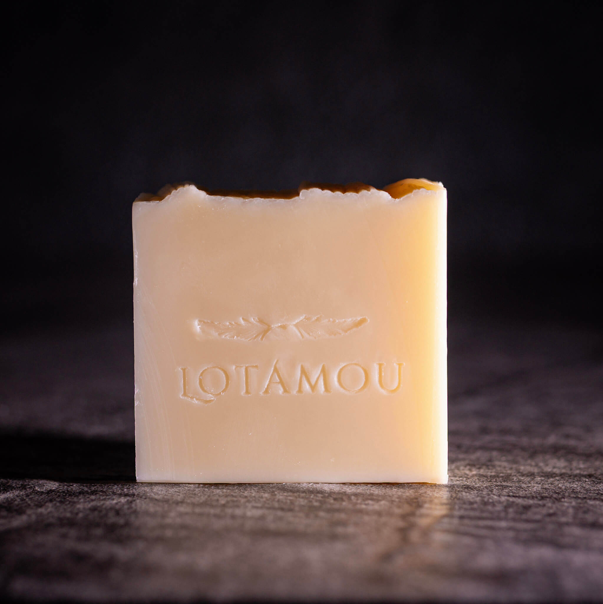 A rectangular bar of Lotamou soap in a warm cream color, embossed with the brand name and a leaf motif, set against a dark, moody background.