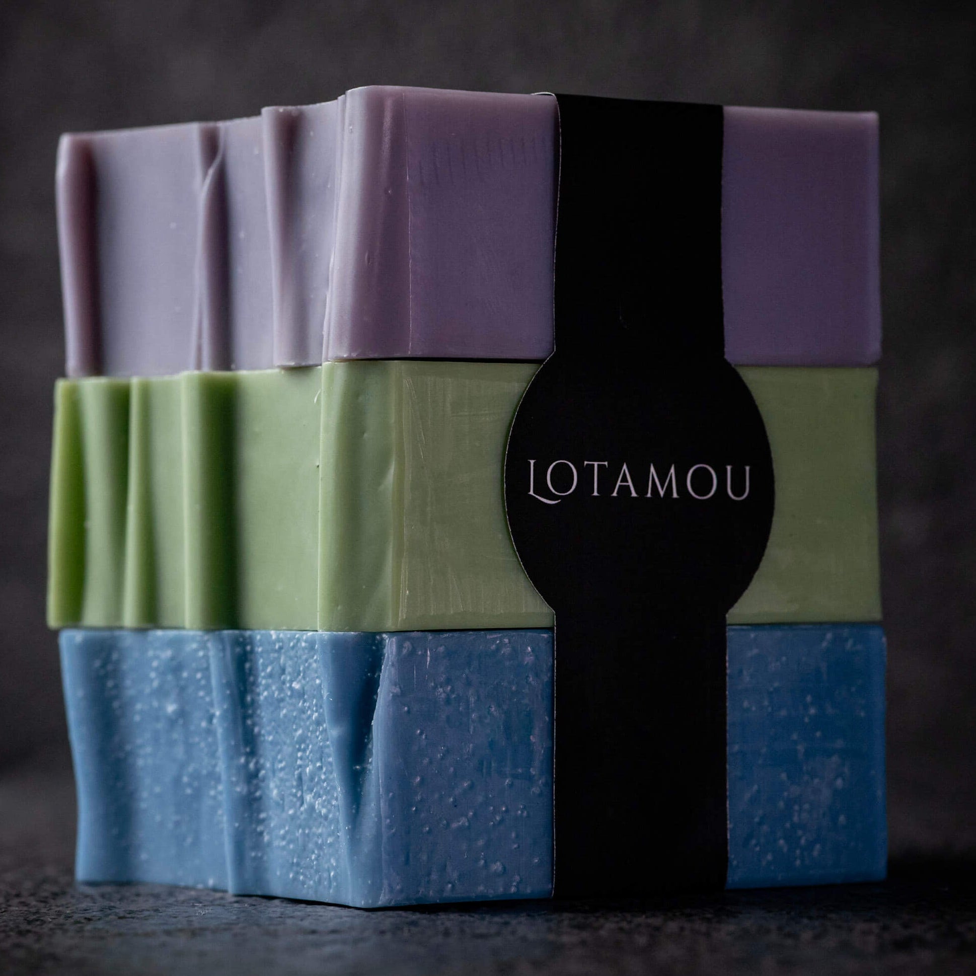 Three stacked rectangular bars of soap in lavender purple, light green, and dusty blue, wrapped together with a black paper band printed with the brand name LOTAMOU.