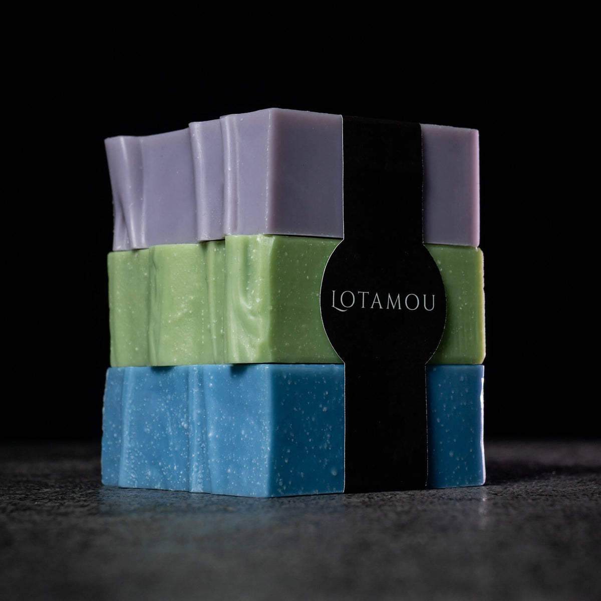 Three stacked rectangular bars of Lotamou soap in lavender, light green, and sky blue, wrapped together with a black paper band, set against a dark background.