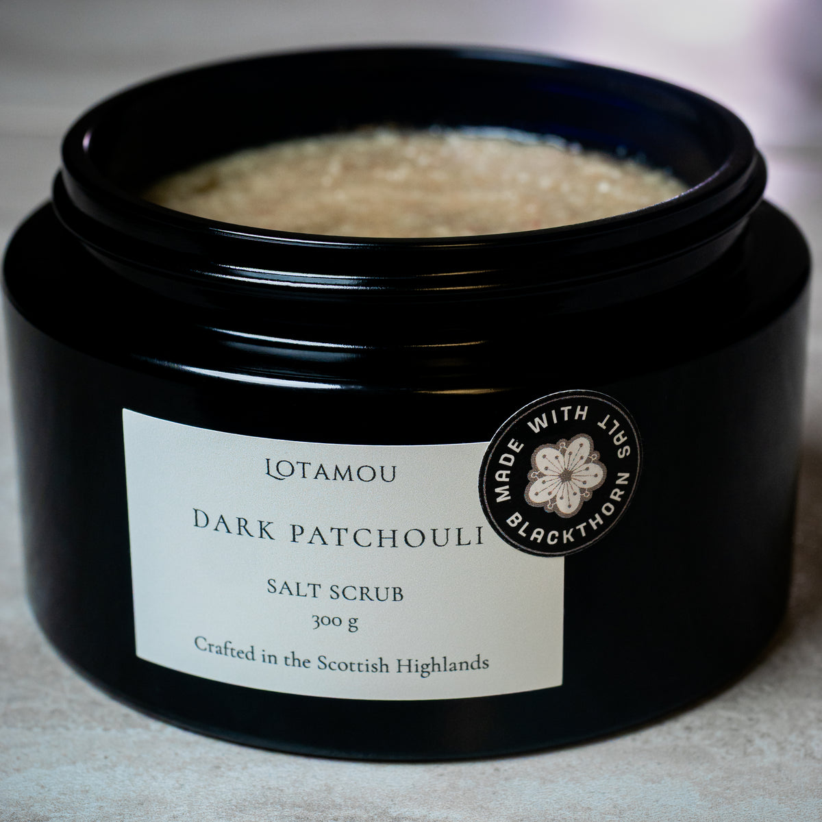A black jar of Lotamou Dark Patchouli Salt Scrub with a white label, shown open to reveal the beige salt scrub inside. A circular 'Made with Love by Blackthorn' badge is on the front right.