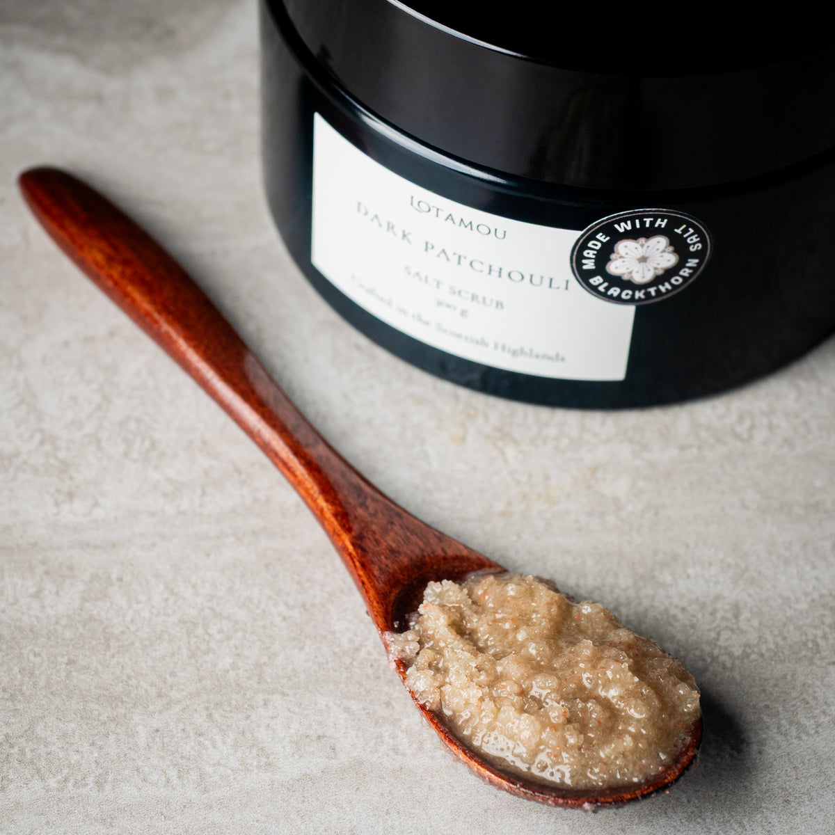 A close-up of a dark jar labeled Lotamou Dark Patchouli Salt Scrub next to a wooden spoon filled with the beige salt scrub, on a light stone surface.