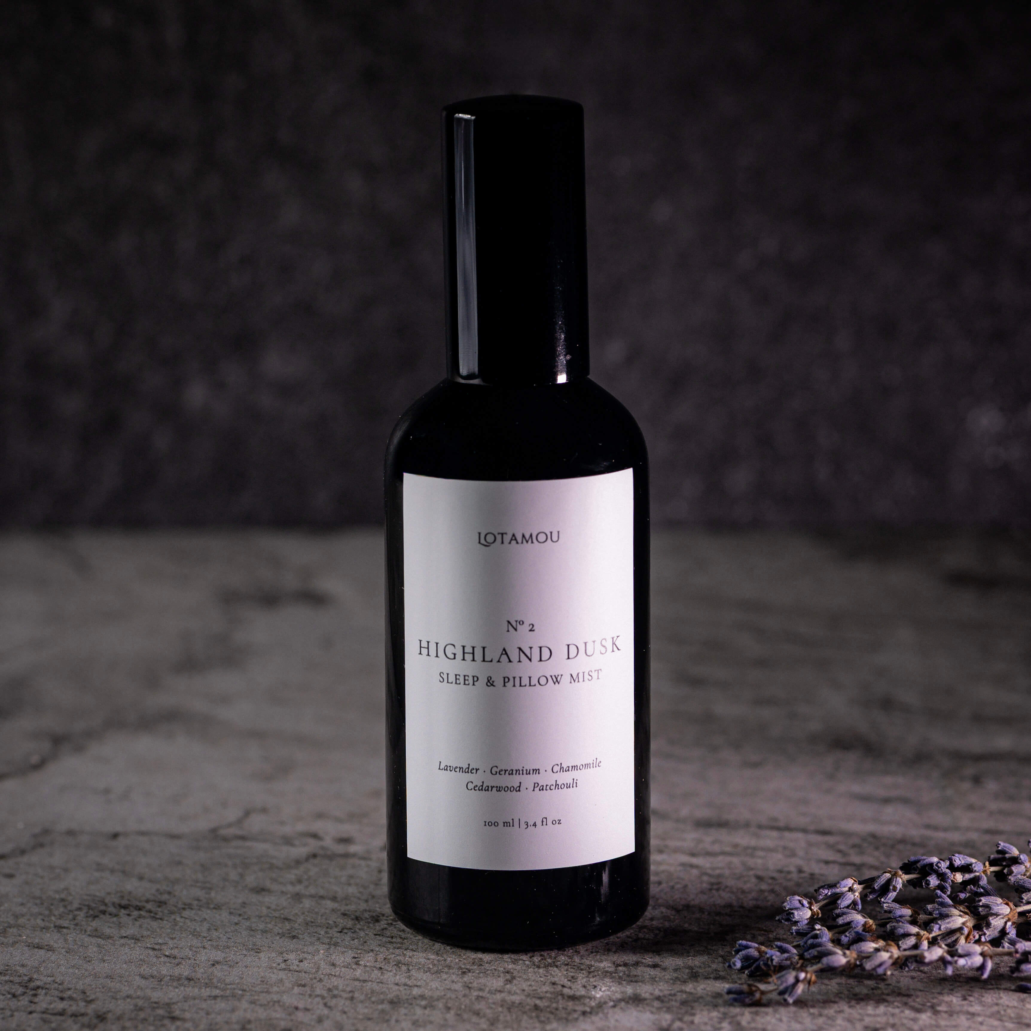 A dark glass bottle of Lotamou 'No. 4 Highland Dusk' sleep and pillow mist, featuring a white label and a black cap, set on a textured gray surface with dried lavender sprigs in the foreground.